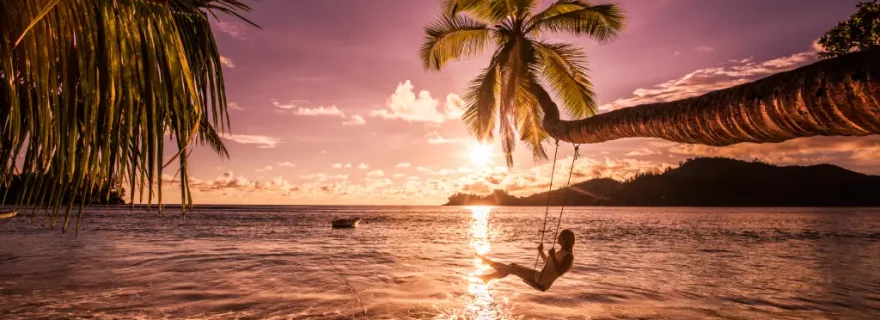 Junge Frau genießt den wunderschönen Sonnenuntergang am Strand, während sie über dem Wasser an einer Palme schaukelt