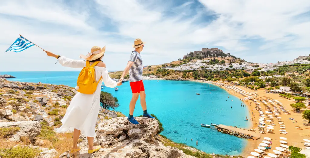 Ein Paar mit einer griechischen Flagge auf einem Hügel mit Blick auf die Bucht und die antike Stadt Lindos auf der Insel Rhodos in Griechenland