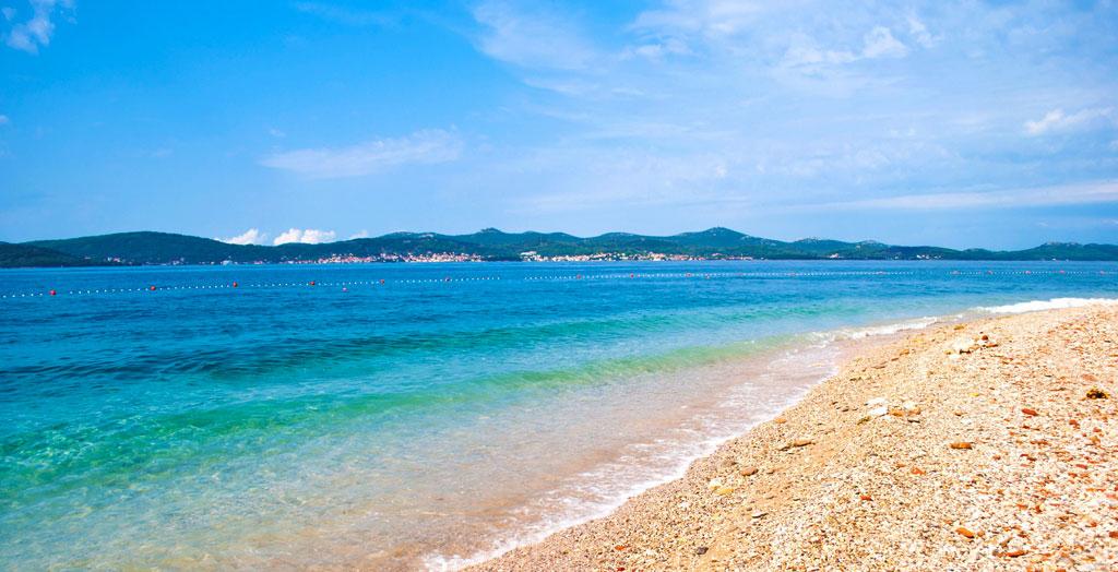 Dieses eindrucksvolle Luftbild präsentiert den Strand von Zadar, eine Küstenperle Kroatiens, in all seiner Pracht. Das azurblaue Meer umschließt sanft die weißen Sandstrände, während die üppigen grünen Bäume eine natürliche Grenze zur urbanen Bebauung bilden. Der idyllische Anblick der friedvollen Bucht vor der lebendigen Kulisse der Stadt Zadar verleiht diesem Ort eine einzigartige Atmosphäre, die sowohl zur Entspannung am Strand als auch zur Erkundung der lokalen Kultur einlädt. Die weitläufige Aussicht lässt erkennen, wie die Natur und städtische Strukturen in perfekter Harmonie koexistieren und unterstreicht die natürliche Schönheit der dalmatinischen Küste.