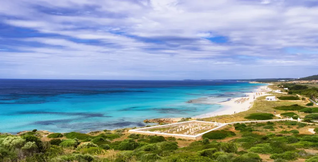 Panoramaansicht des Strandes von Son Bou auf Menorca, umrahmt von grüner Landschaft und azurblauem Wasser