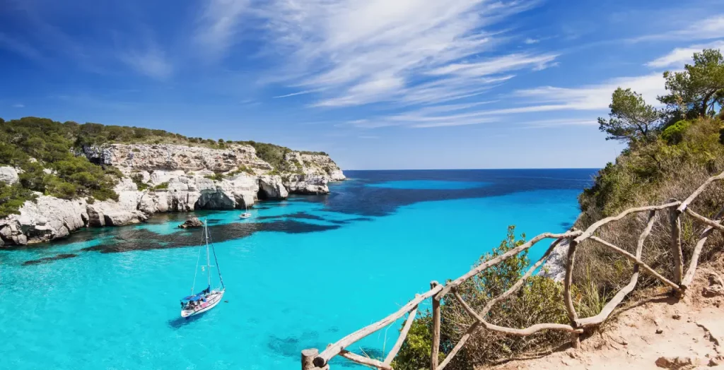 Atemberaubende Aussicht auf eine abgelegene Bucht auf Menorca mit klarem türkisfarbenem Wasser und traditionellem Segelboot