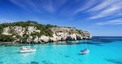 Zwei Segelboote ankern in der einsamen türkisfarbenen Bucht von Menorca umgeben von felsiger Küste und grünen Bäumen