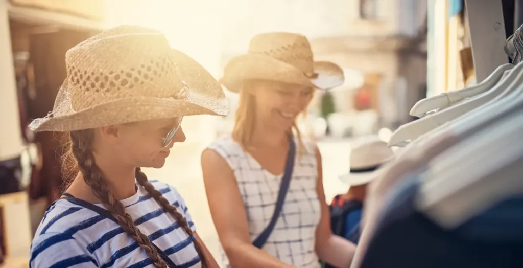 Zwei Frauen in Sommerkleidung und Strohhüten beim Einkaufen in einer sonnigen Urlaubsstraße