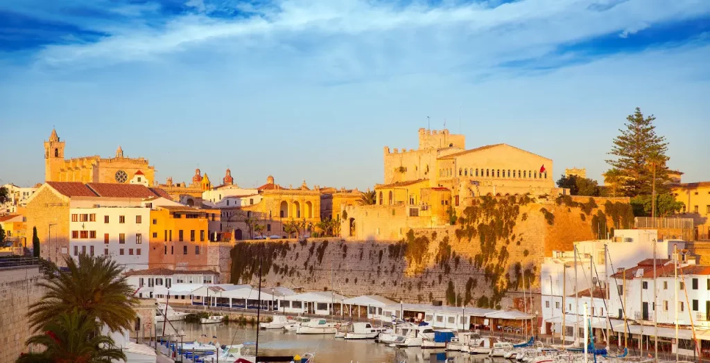 Historischer Hafen von Ciutadella bei Sonnenuntergang mit Gebäuden aus Marès-Sandstein und ruhigen Gewässern auf Menorca [Bildquelle: © Lunamarina | Canva]