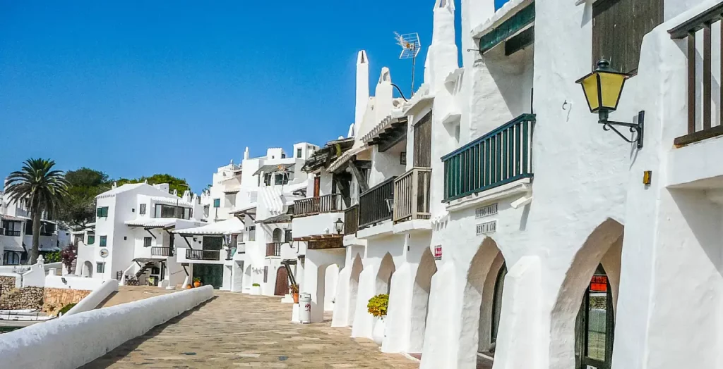 Straße in Menorca mit historischen weißen Häusern und Bögen, traditionellen Balkonen und Straßenlaternen [Bildquelle: © Scrisman| Canva]