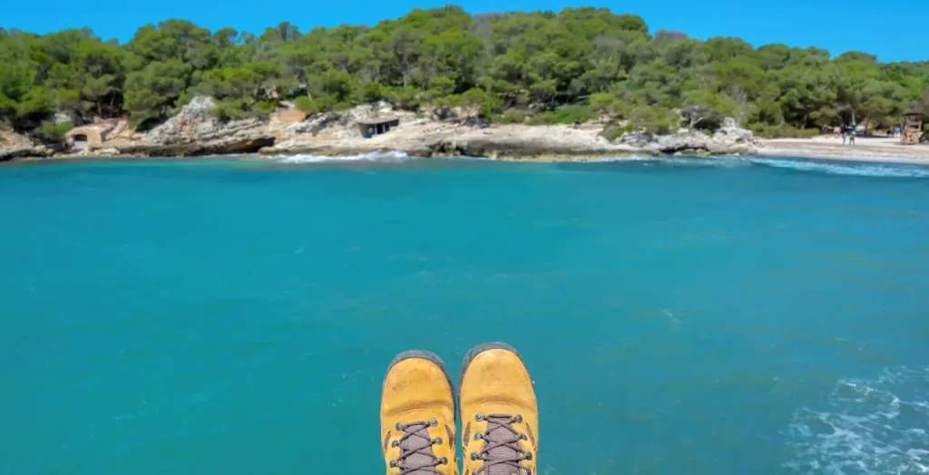 Perspektive eines Wanderers über türkisfarbenem Meer an der Küste Menorcas, mit Wanderstiefeln im Vordergrund