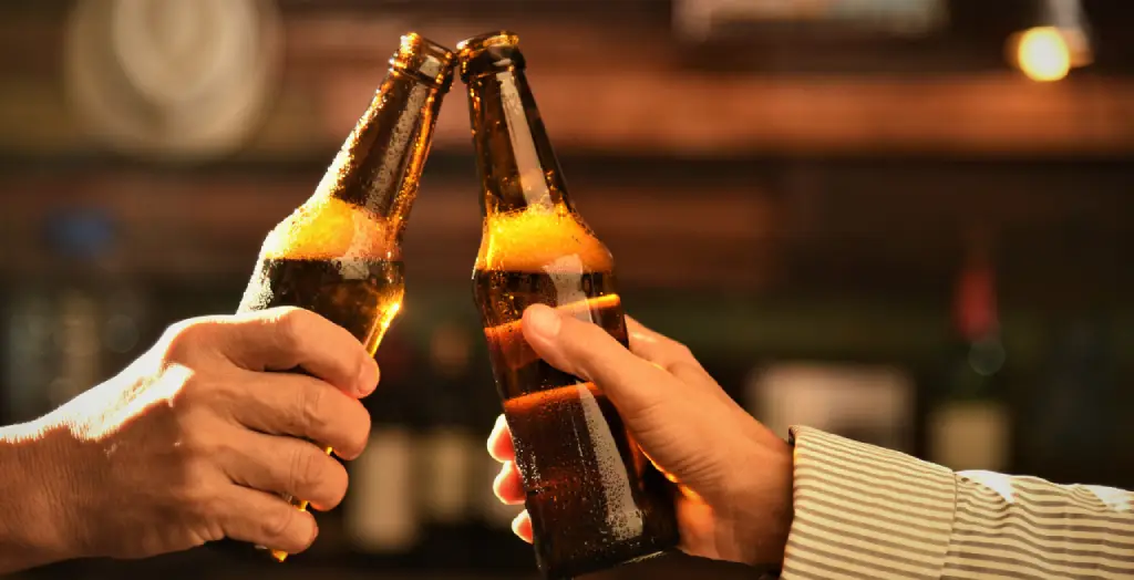 Zwei Hände stoßen mit Bierflaschen an, spritzende Biertröpfchen im warmen Licht