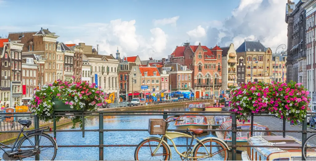 Fahrrad geparkt am Geländer einer Gracht in Amsterdam, Niederlande, mit blühenden Blumen und historischen Gebäuden im Hintergrund [Bildquelle: © tunart | Canva]