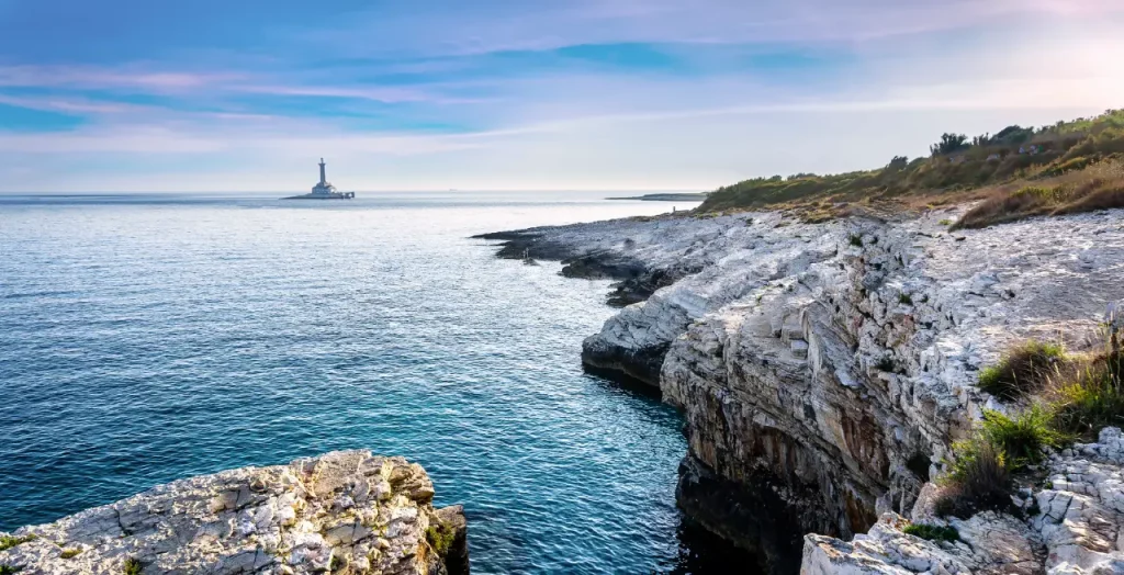 Blick auf das ruhige Meer vom Kap Kamenjak in Kroatien mit einem Leuchtturm im Hintergrund und felsiger Küstenlinie im Vordergrund [Bildquelle: © Wirestock | Canva]