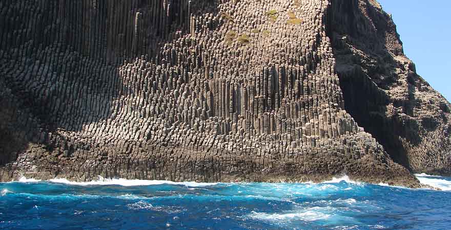 Felswand die aussieht wie Orgelpfeifen an der sich die Wellen brechen.
