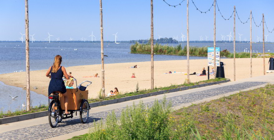 Eine Frau mit blauem Kleid fährt mit Kind (links im Bild) im Sommer auf einem Lastenfahrrad auf dem Radweg entlang des Vesuwemeers. Zu sehen ist auch ein Strandabschnitt mit einer Hand voll sich sonnenden Menschen sowie Windräder im Hintergrund. 