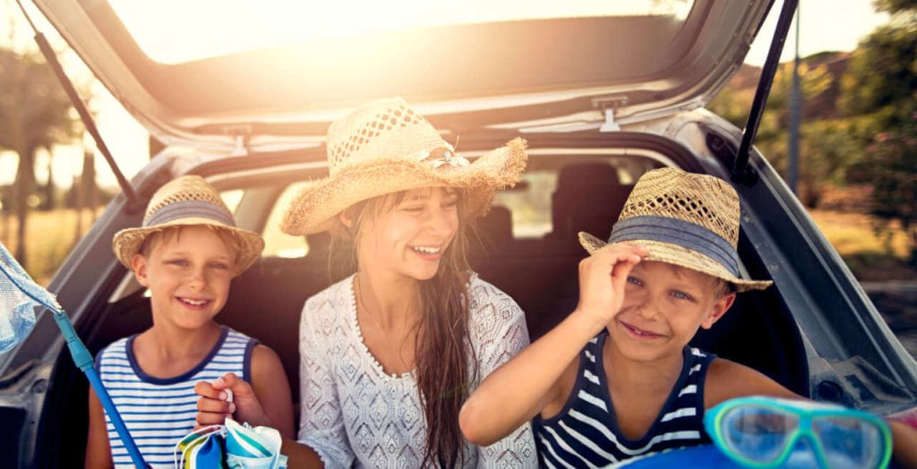 Drei Kinder sitzen mit Sonnenhut im Kofferraum eines Autos