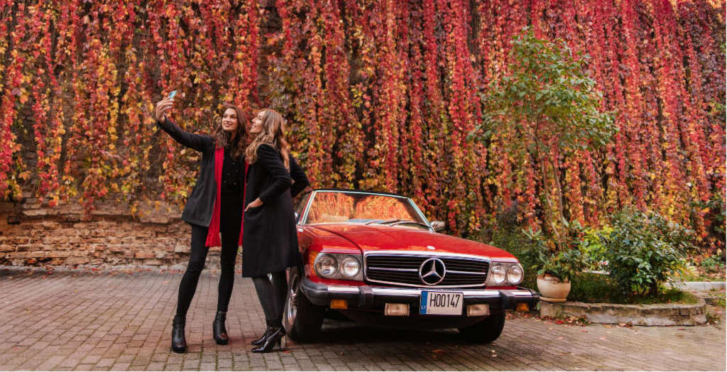 zwei Frauen nehmen ein Selfie auf im Hintergrund ein rotes Auto und ein blühender Baum in Vilnius