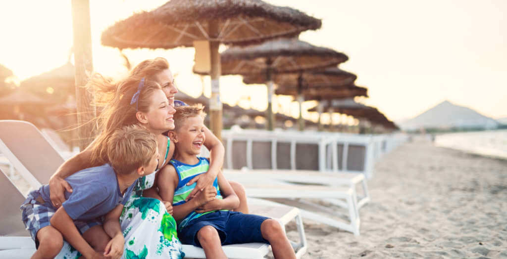 Frau sitzt mit ihren Kindern am Strand unterm Sonnenschirm