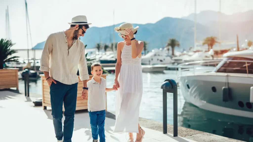 Stilvolle Familie schlendert Hand in Hand durch den Hafen von Porto Montenegro, umgeben von Luxusyachten und malerischen Bergsilhouetten