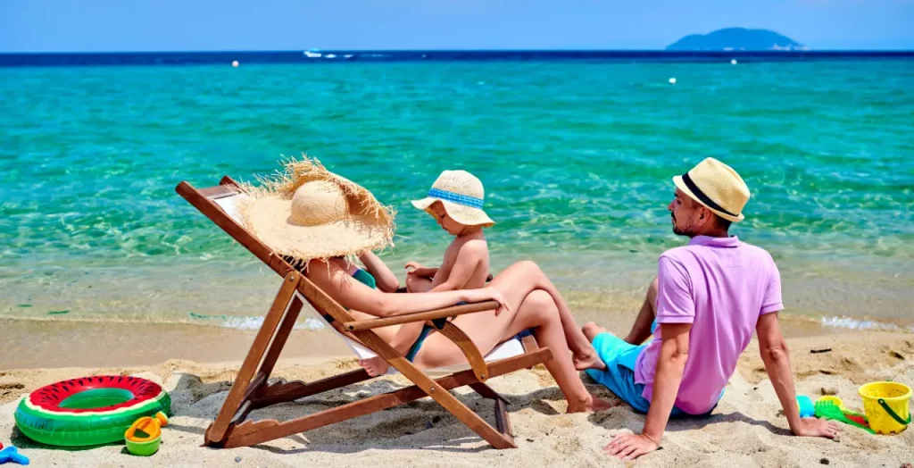 Familie entspannt auf Liegestühlen am Strand von Griechenland, umgeben von klarem blauem Wasser [Bildquelle: © haveseen | Canva]
