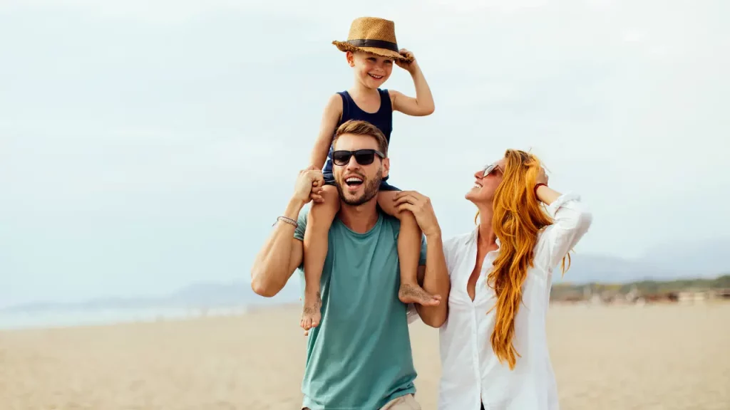 Junge Familie genießt einen entspannten Tag am Strand von Montenegro, Kind auf Vaters Schultern und Mutter lachend daneben [Bildquelle: © mihailomilovanovic | Canva]