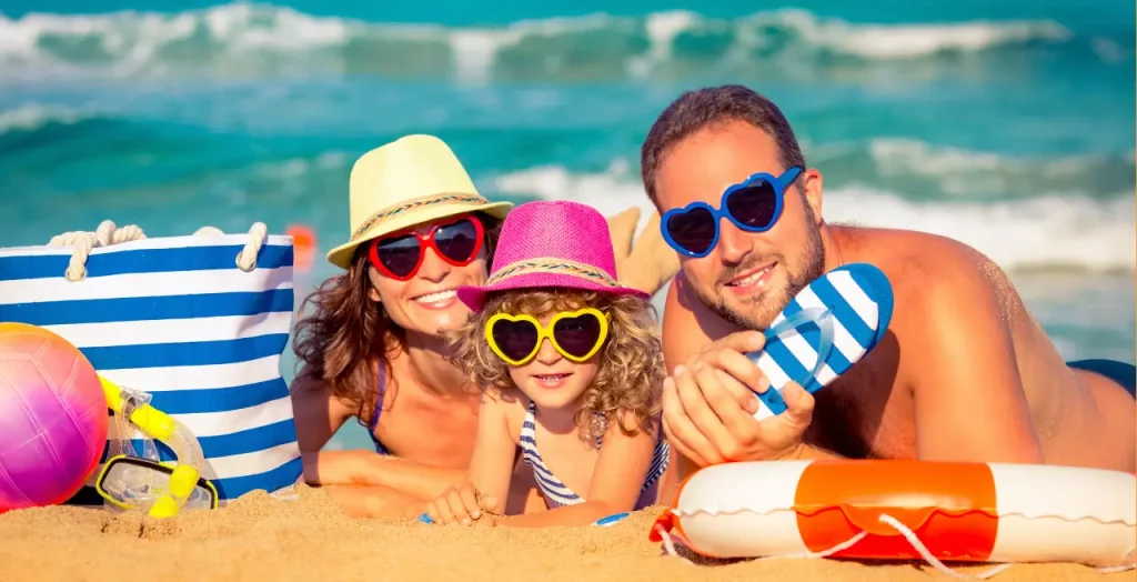Fröhliche Familie mit Sonnenhüten und herzförmigen Sonnenbrillen entspannt am Strand [Bildquelle: © yaruta  | Canva]