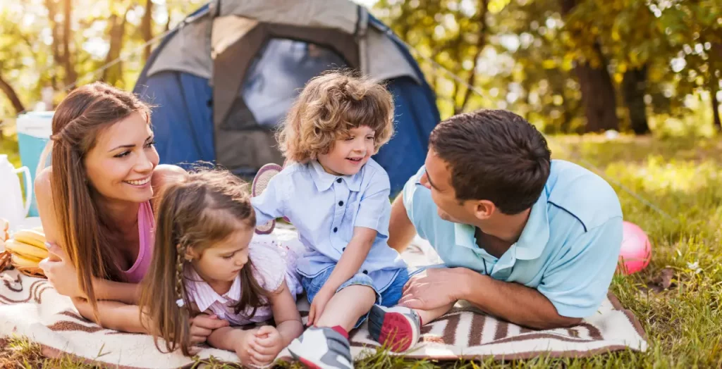Glückliche Familie beim Camping vor einem Zelt, entspannend auf einer Decke im Wald [Bildquelle: © skynesher | Canva]