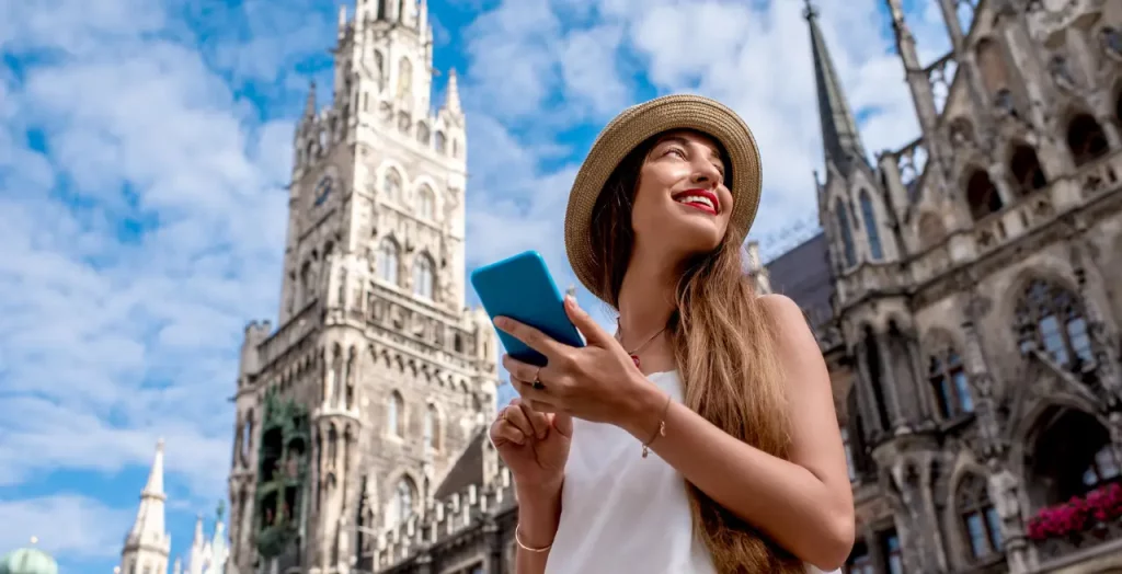 Junge Frau mit Strohhut benutzt Smartphone vor dem Münchener Rathaus in Deutschland [Bildquelle: © RossHelen | Canva]