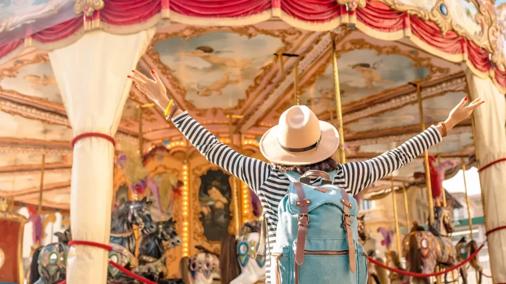 Frau mit Hut und gestreiftem Oberteil hebt Arme vor einem historischen Karussell [Bildquelle: © frantic00  | Canva]