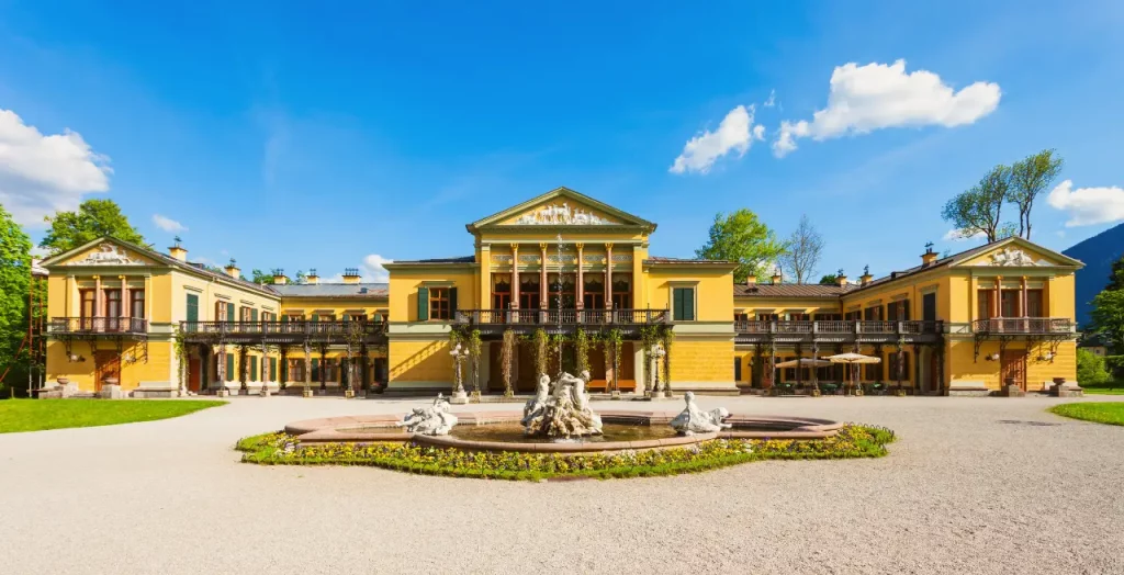 Die Kaiservilla in Bad Ischl, Österreich, mit ihrem eleganten gelben Anstrich und umgebenden Gärten unter blauem Himmel [Bildquelle: © Andrey Khrobostov | Canva]