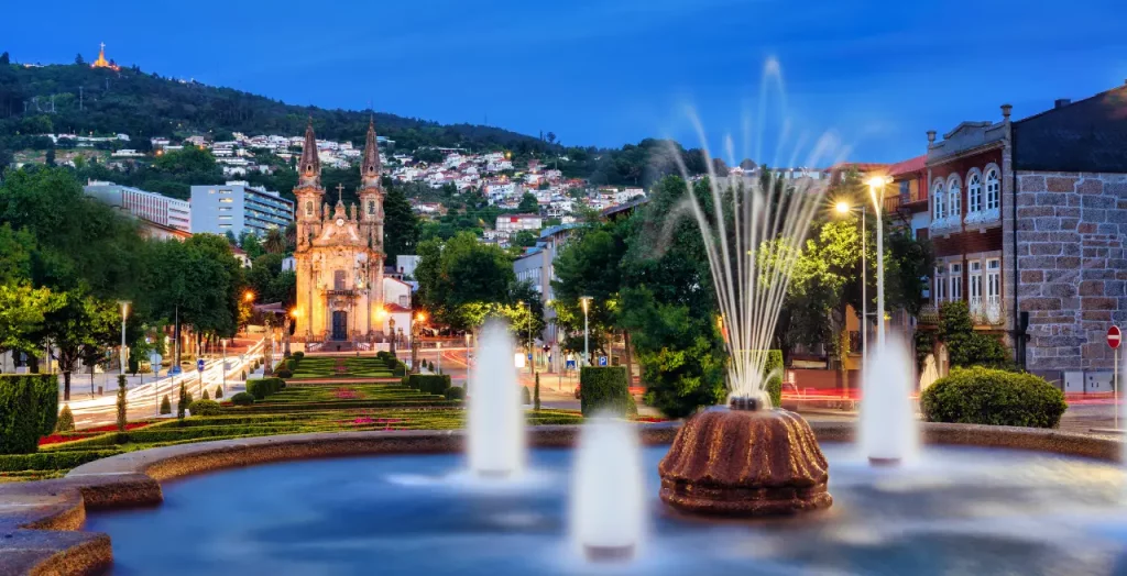 Nächtliche Ansicht des Stadtzentrums von Guimarães, Portugal, mit beleuchteter Kirche und Brunnen im Vordergrund [Bildquelle: © Armando Oliveira | Canva]