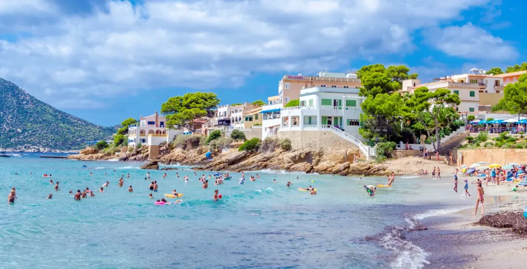 Sommerliche Stimmung am Strand Sant Elm mit badenden Urlaubern und malerischen Häusern auf Mallorca, Spanien